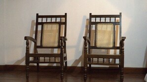 Twee houten stoelen met geweven rugleuning tegen een effen muur.