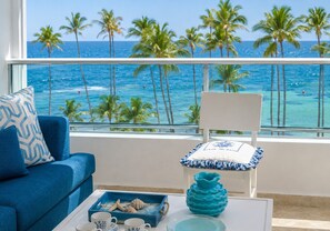 A balcony with a view of the ocean and palm trees, a blue sofa, a white chair, and decorative items on a table.
