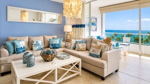 Een moderne woonkamer met een groot raam met uitzicht op de oceaan, een beige bank met blauwe en witte kussens, een witte salontafel en een hanglamp.