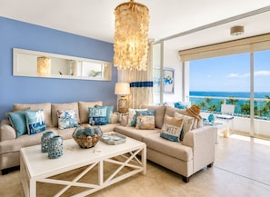 A modern living room with a large window overlooking the ocean, a beige sofa with blue and white cushions, a white coffee table, and a hanging light fixture.