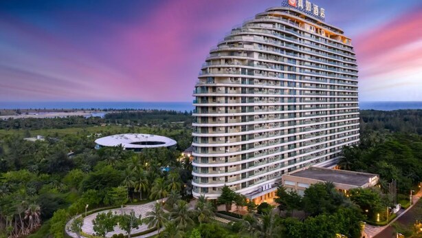 A large, multi-story hotel with a curved facade, surrounded by lush greenery and a view of the ocean. - Qionghai Bozha Baiyuewan Zhenru Hotel (Qionghai)
