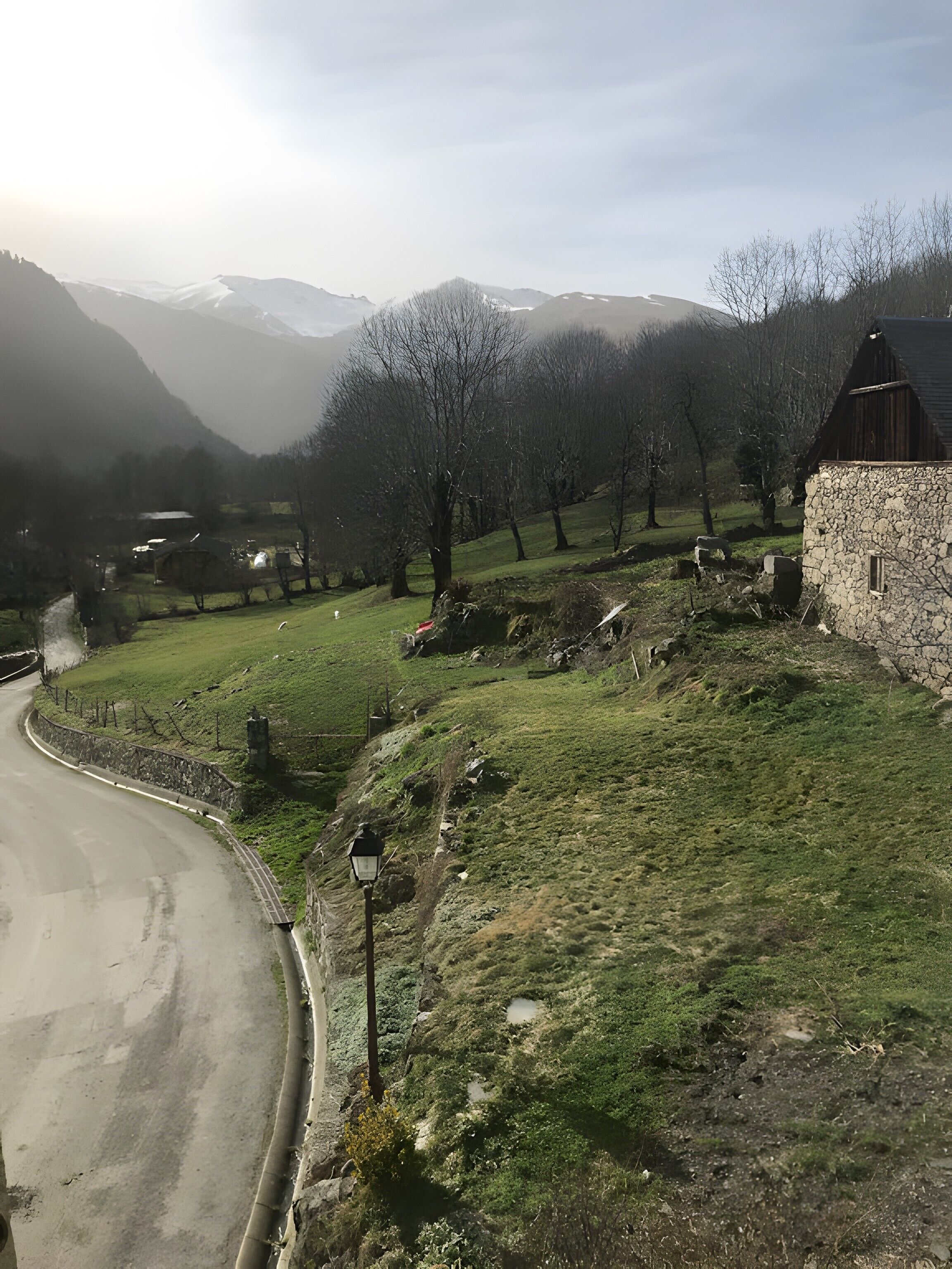 Un paysage rural avec un bâtiment en pierre, une route sinueuse et une chaîne de montagnes en arrière-plan.