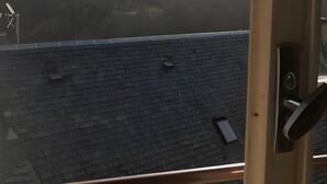 Une vue à travers la fenêtre d’un bâtiment doté d’un toit en tuiles, d’une rambarde en métal et, au loin, d’un paysage montagneux.