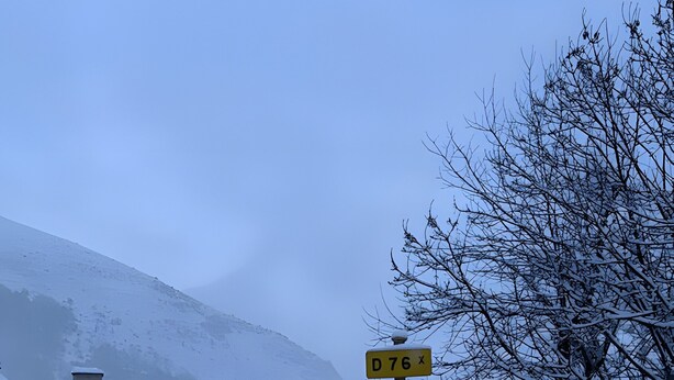 Une scène hivernale représentant un village enneigé, avec un panneau indiquant « CAZEAUX DE LARBOUST » et un indicateur de distance indiquant « 0,76 km ».