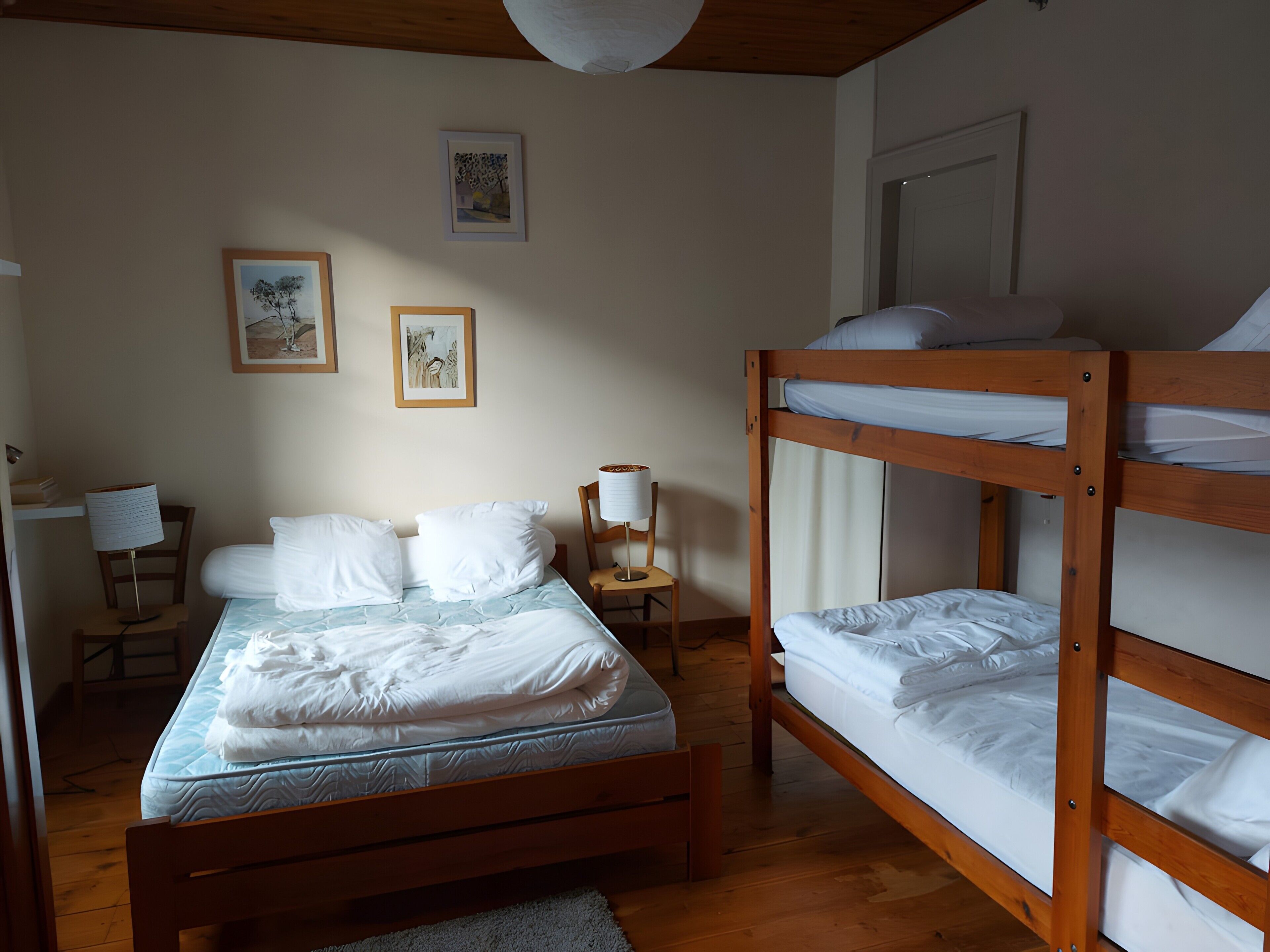A room with a bunk bed, a single bed, a chair, a lamp, and two framed pictures on the wall.