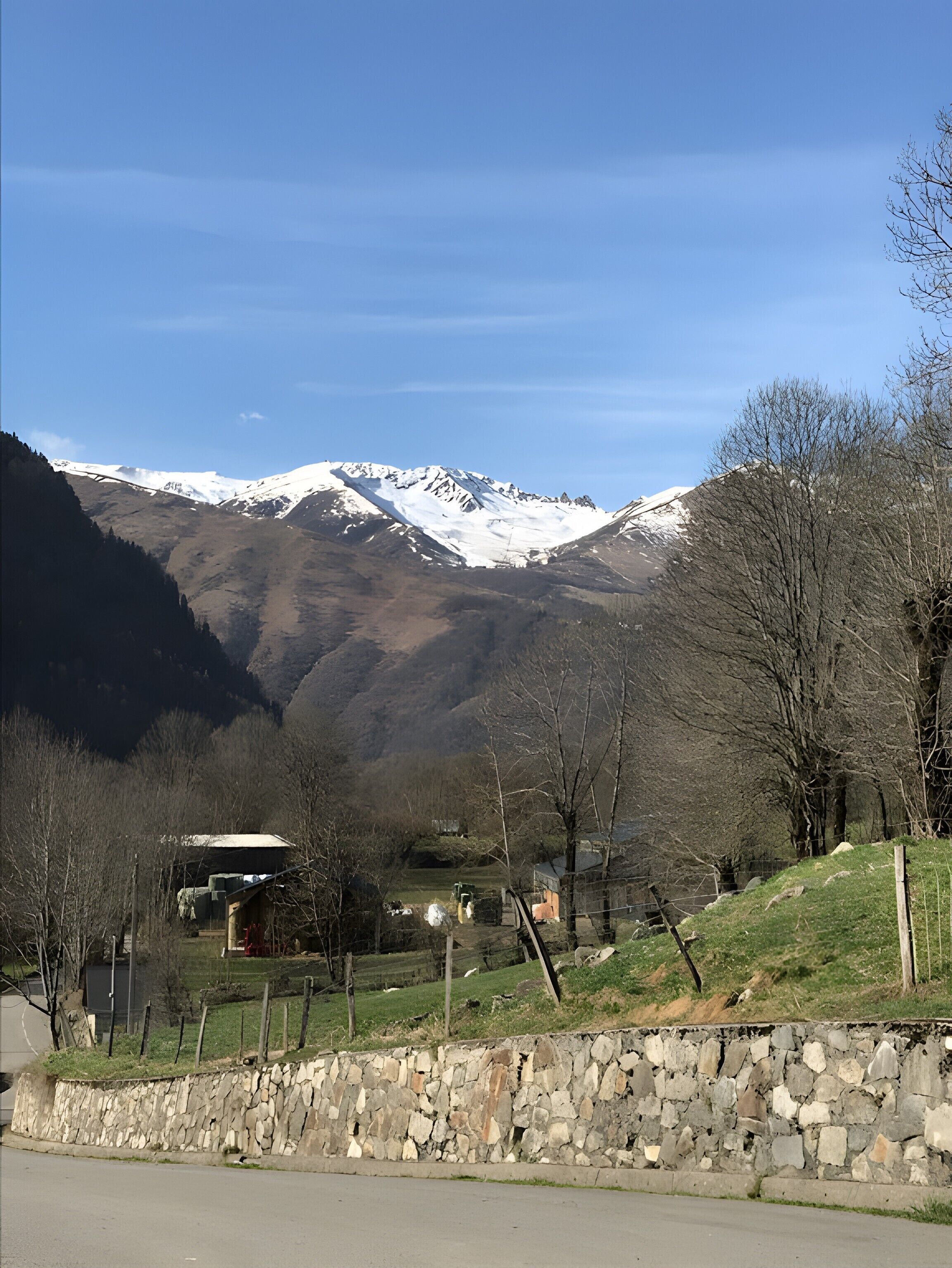 A rural landscape with a stone wall, a building, and snow-capped mountains in the background.