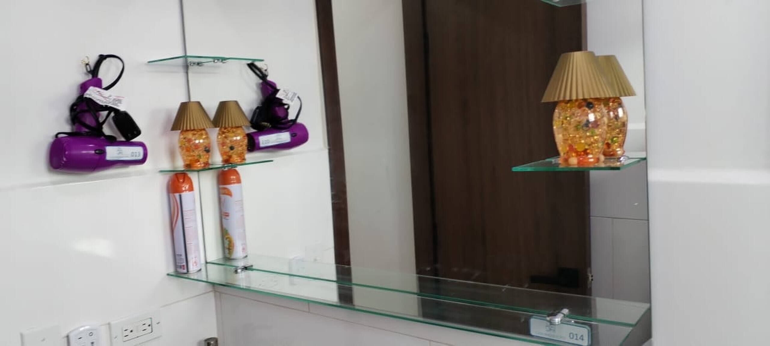 Un bagno con ripiano in vetro che contiene una lampada e un oggetto decorativo. Si notano anche due bottiglie viola e una bottiglia bianca a parete.