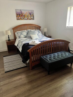 Quarto com cama de madeira, um banquinho, mesa de cabeceira, luminária e um quadro na parede.