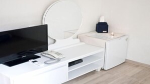 A modern hotel room with a flat-screen TV, a bedside table with a phone, a small heater, and a compact refrigerator.