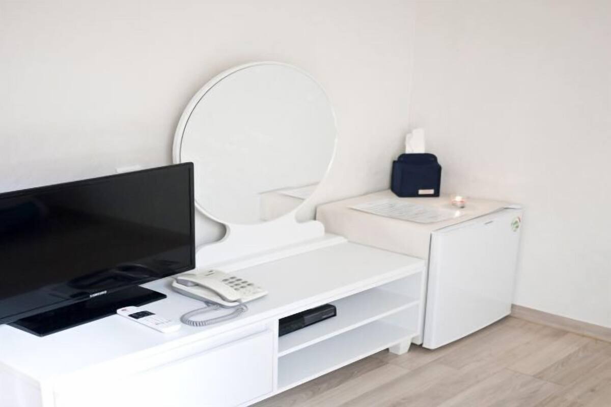 A modern hotel room with a flat-screen TV, a bedside table with a phone, a small heater, and a compact refrigerator.