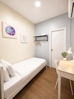 Habitación individual con cama, piso de madera, un escritorio blanco con lámpara, estante colgante y dos carteles con mensajes motivacionales en la pared.