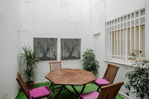 Una mesa de madera hexagonal rodeada de cuatro sillas, dos cuadros colgados en la pared, una planta en maceta y una ventana con barrotes blancos.