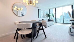 A modern living room with a round dining table, a sofa, and a large window with a city view. - Luxury apartment & free parking x1 (Southbank)