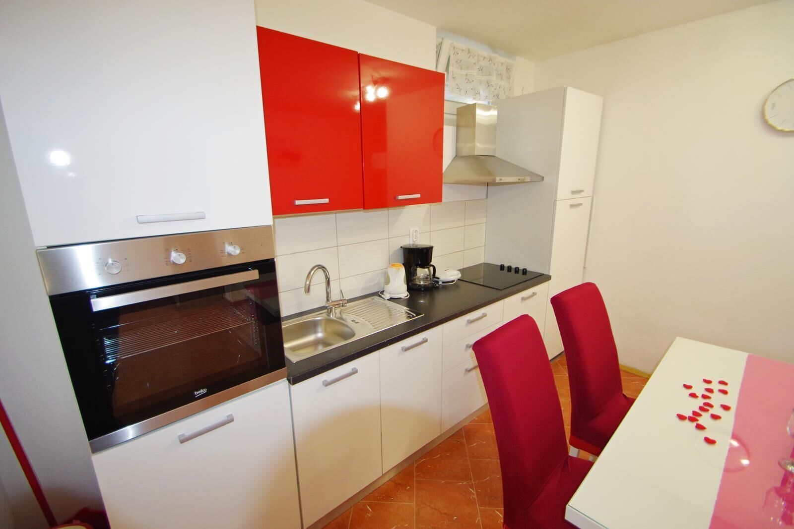 Une cuisine compacte avec des meubles blancs et rouges, un four noir, un évier en acier inoxydable et une table avec des chaises rouges.