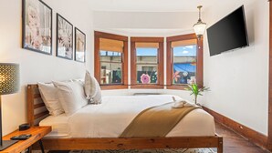 Un dormitorio con una cama grande, un televisor montado en la pared y una ventana con vistas a edificios.