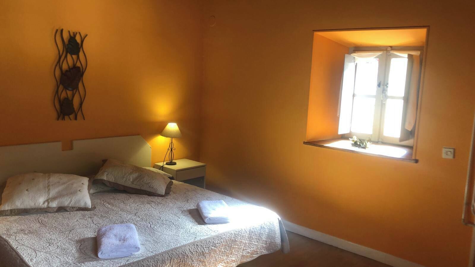 Et soveværelse med en seng, puder, et natbord med en lampe og et vindue med gardiner.