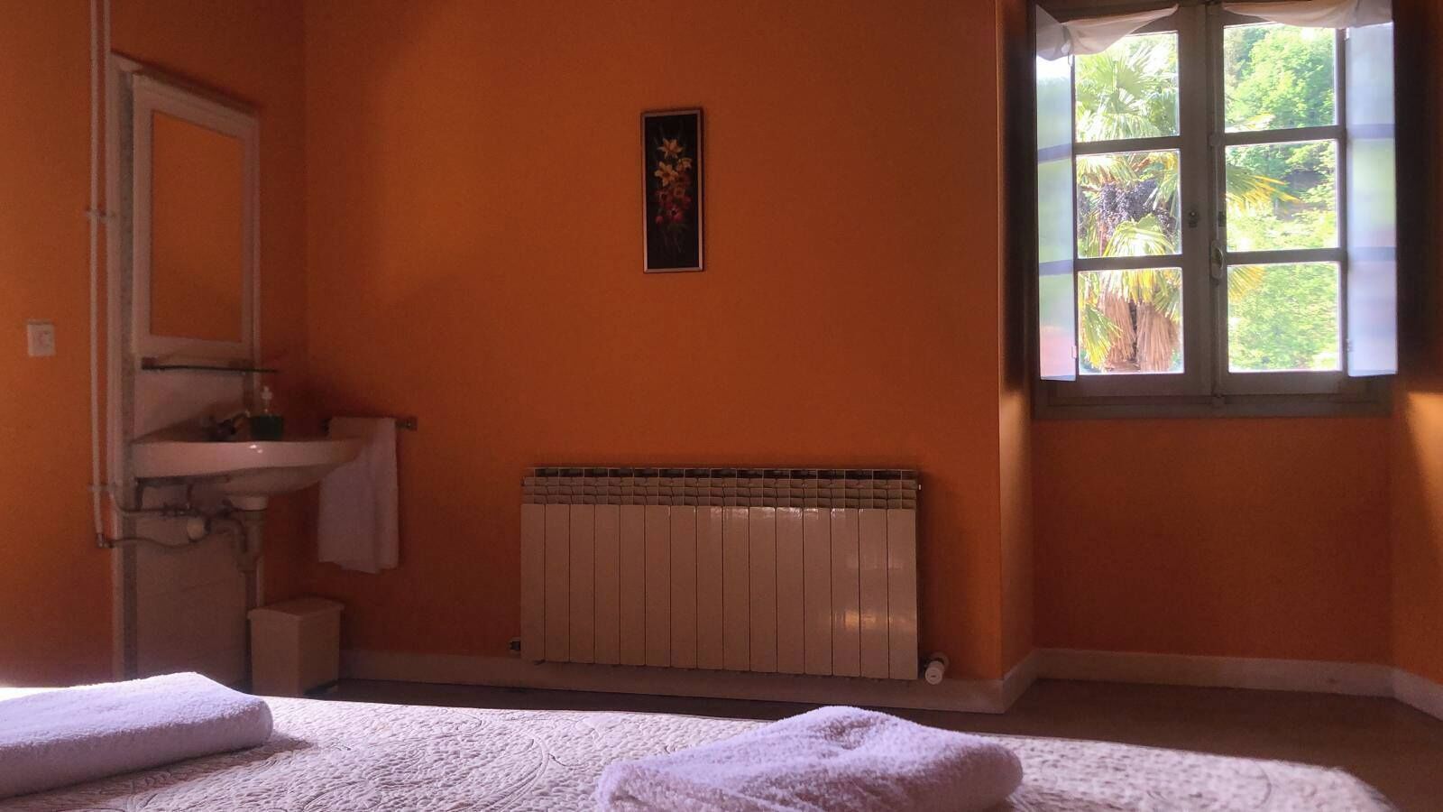 A room with an orange wall, a radiator, a window with a view of greenery, a sink, and a mirror.