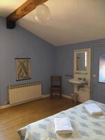 Un dormitorio con techo de madera, un radiador, una silla, una cama con colcha estampada, un lavamanos blanco y un cuadro en la pared.
