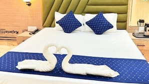 A hotel room with a bed, pillows, a wall-mounted air conditioner, and a nightstand with a lamp and a coffee maker. - Hotel Mangalam (Varanasi)
