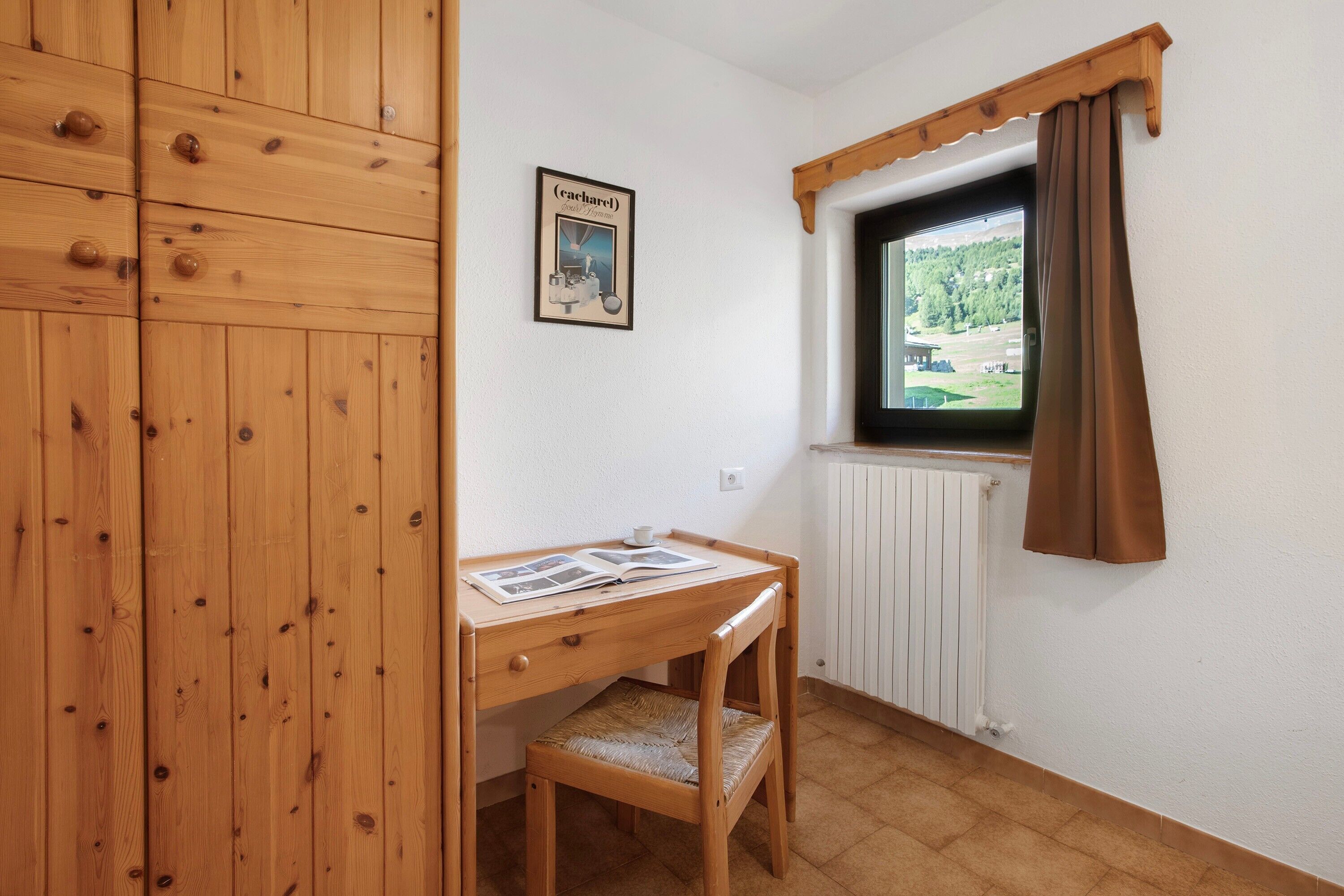 A wooden desk with a chair, a radiator, a window with curtains, and a framed picture on the wall.