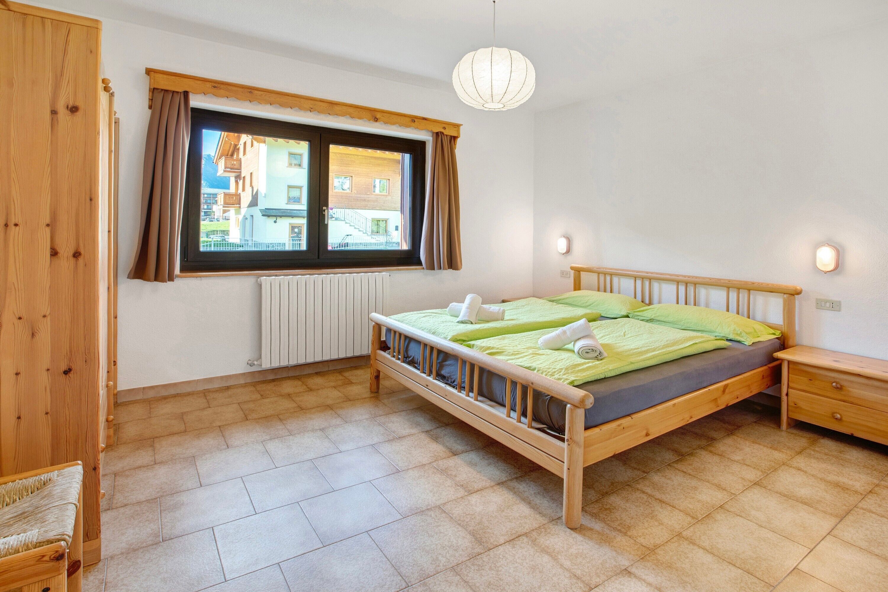 Une pièce avec un lit en bois, une fenêtre avec des rideaux, un radiateur et une table de chevet en bois.
