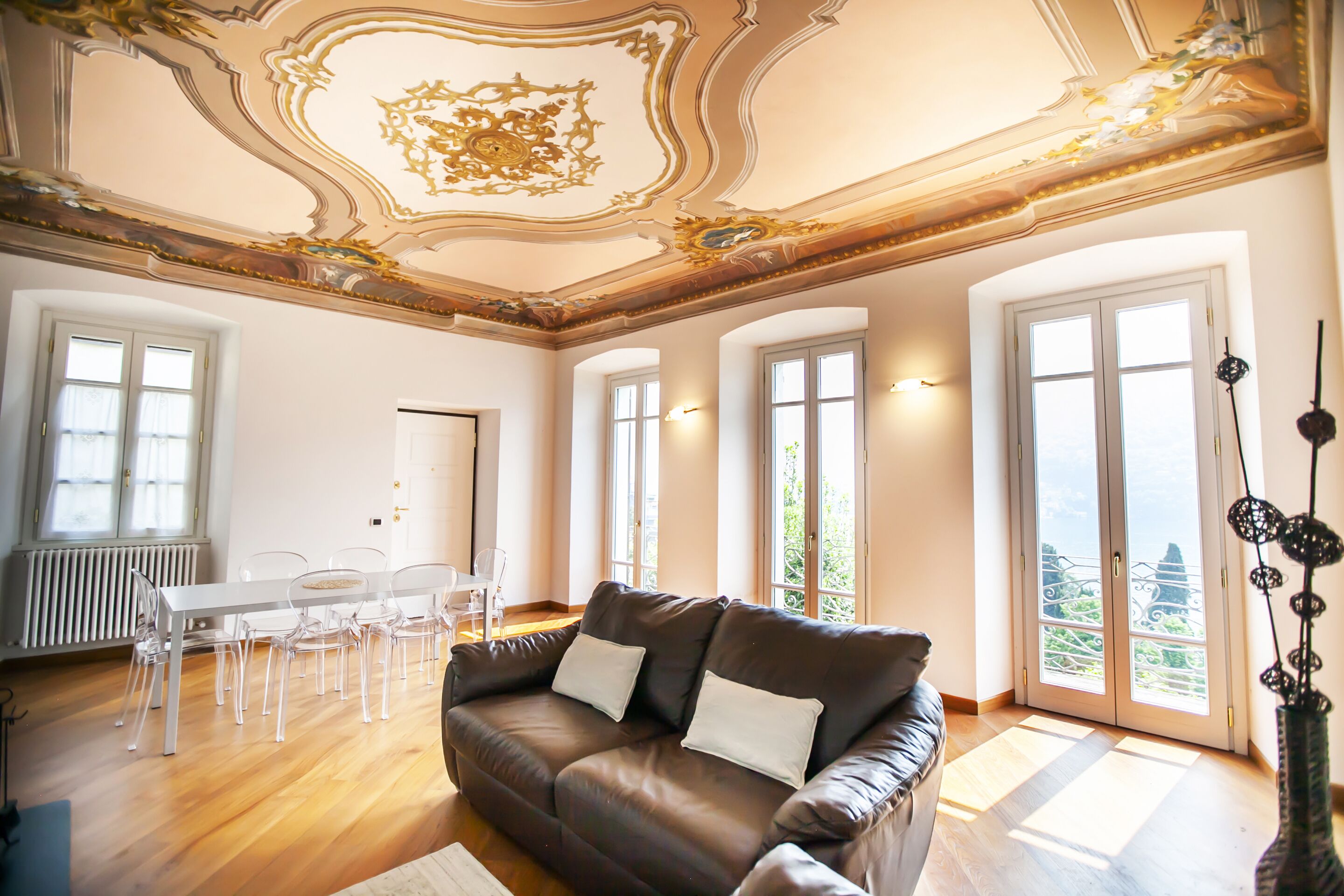 A spacious living room with a brown leather sofa, a white dining table with chairs, and large windows offering a view of greenery.