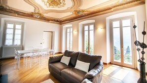 A spacious living room with a brown leather sofa, a white dining table with chairs, and large windows offering a view of greenery. - Appartamento Fioravante by Wonderful Italy (Moltrasio)