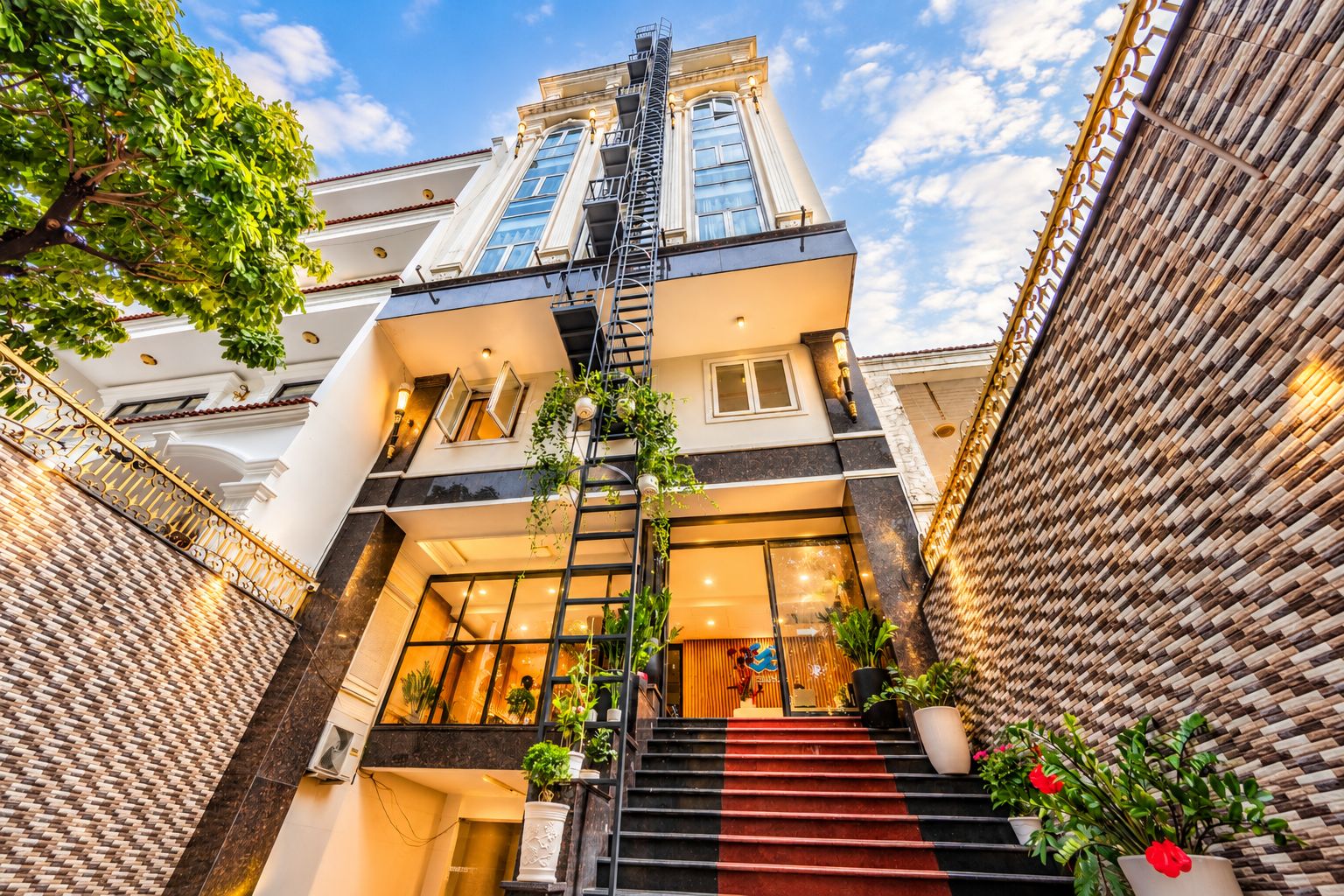 A multi-story building with a staircase leading to the entrance, surrounded by greenery and a woven wall.