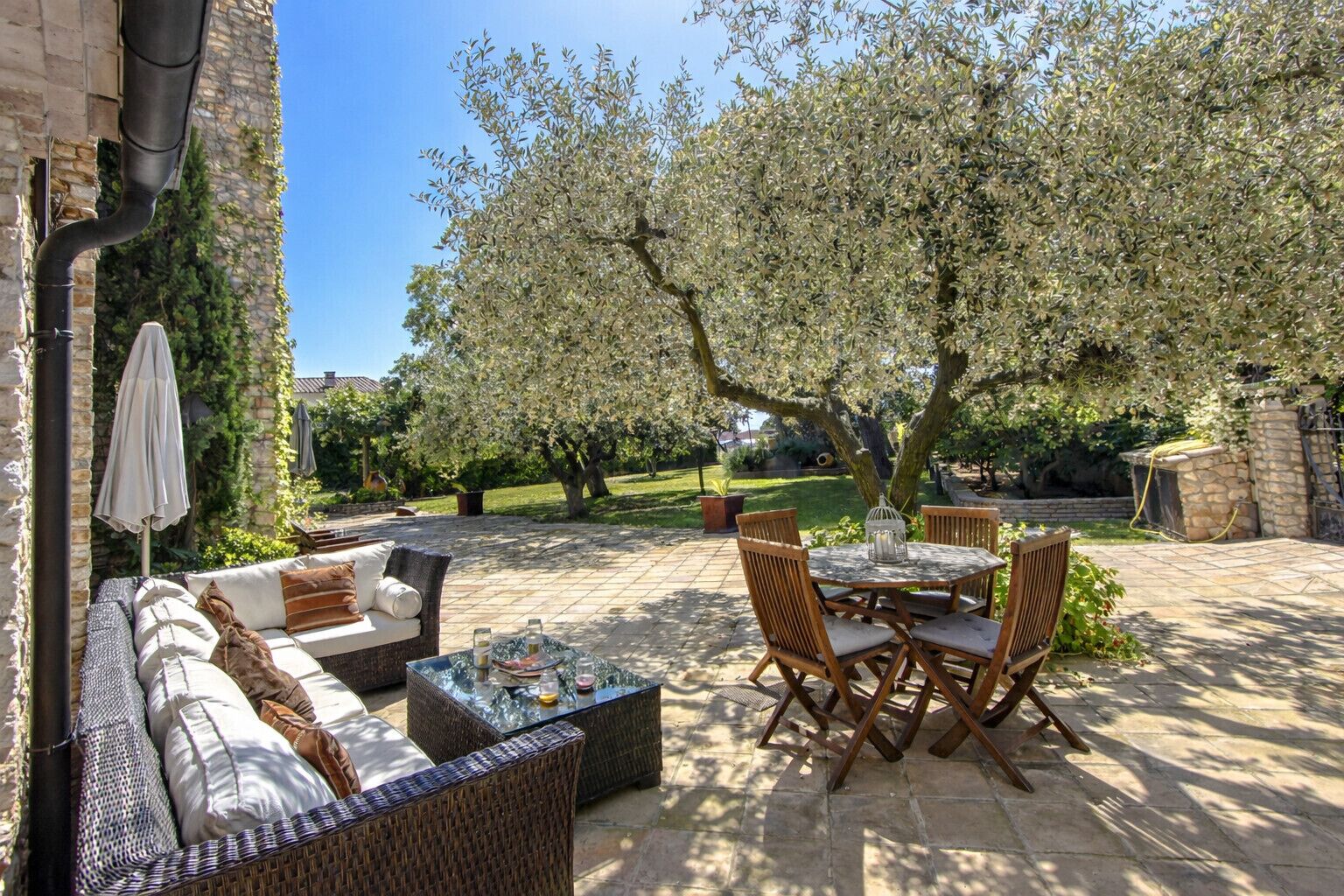 Un espacio de patio al aire libre con un sofá, una mesa de comedor y sillas, rodeado de árboles y un muro de piedra.