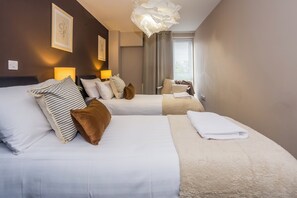 Una habitación de hotel con dos camas, un ventanal con cortinas, una araña y cuadros en las paredes.
