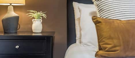 Una cama bien hecha con almohadas blancas y a rayas, una almohada de acento marrón y una lámpara de noche sobre una mesita auxiliar negra.
