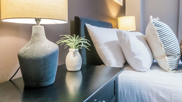 A neatly made bed with white and striped pillows, a vase with a plant, and a lamp on a dark bedside table.