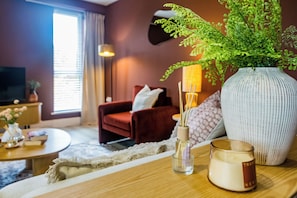 A cozy living room with a red sofa, a large vase with green foliage, a small table with candles, and a television in the background. - Toast Stays - The Ruby at Parsons House (Washington)
