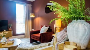 A cozy living room with a red sofa, a large vase with green foliage, a small table with candles, and a television in the background.