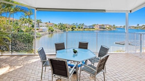 A covered outdoor patio with a glass table and chairs, overlooking a body of water and residential buildings. - Villa Maria 4BRM Home (Clear Island Waters)