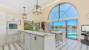 A modern kitchen with a large island, white cabinetry, and a dining area with a view of a pool and resort buildings. - Villa Maria 4BRM Home (Clear Island Waters)