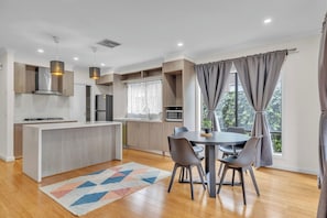 A modern kitchen with a central island, dining table with chairs, and a view of greenery outside.