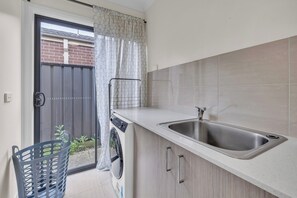 A kitchen with a stainless steel sink, a washing machine, and a small window with a curtain.