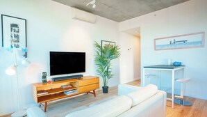 Uma sala de estar moderna com uma televisão de tela plana, um móvel de entretenimento de madeira, um sofá branco, uma área de jantar com uma mesa pequena e cadeiras, e uma planta em vaso.