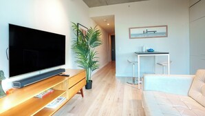 Uma sala de estar moderna com televisão de tela plana, painel de entretenimento de madeira, planta em vaso e um espaço de jantar com mesa pequena e banqueta.