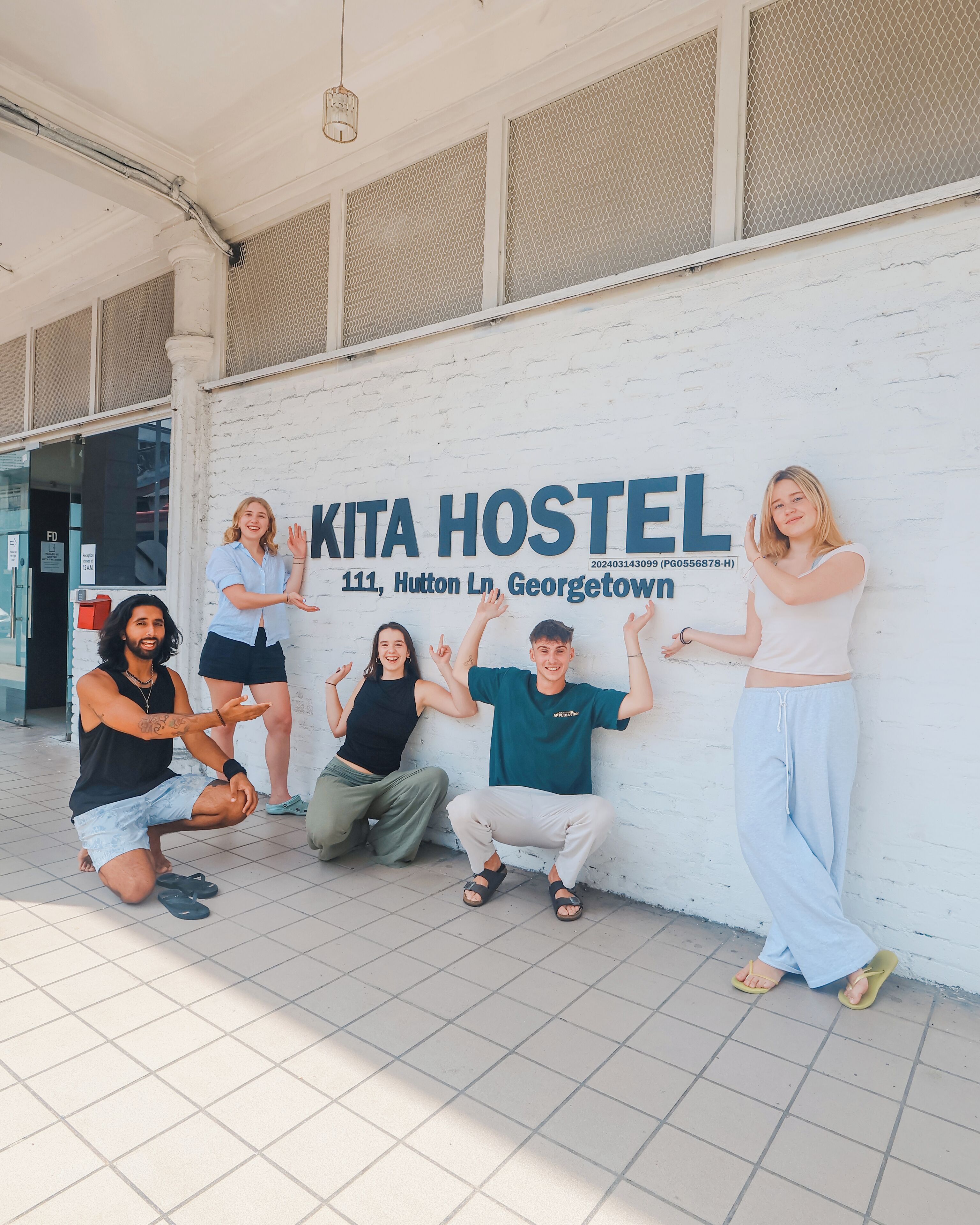 A group of people posing in front of a hostel named "KITA HOSTEL" with a sign displaying the address and contact number.