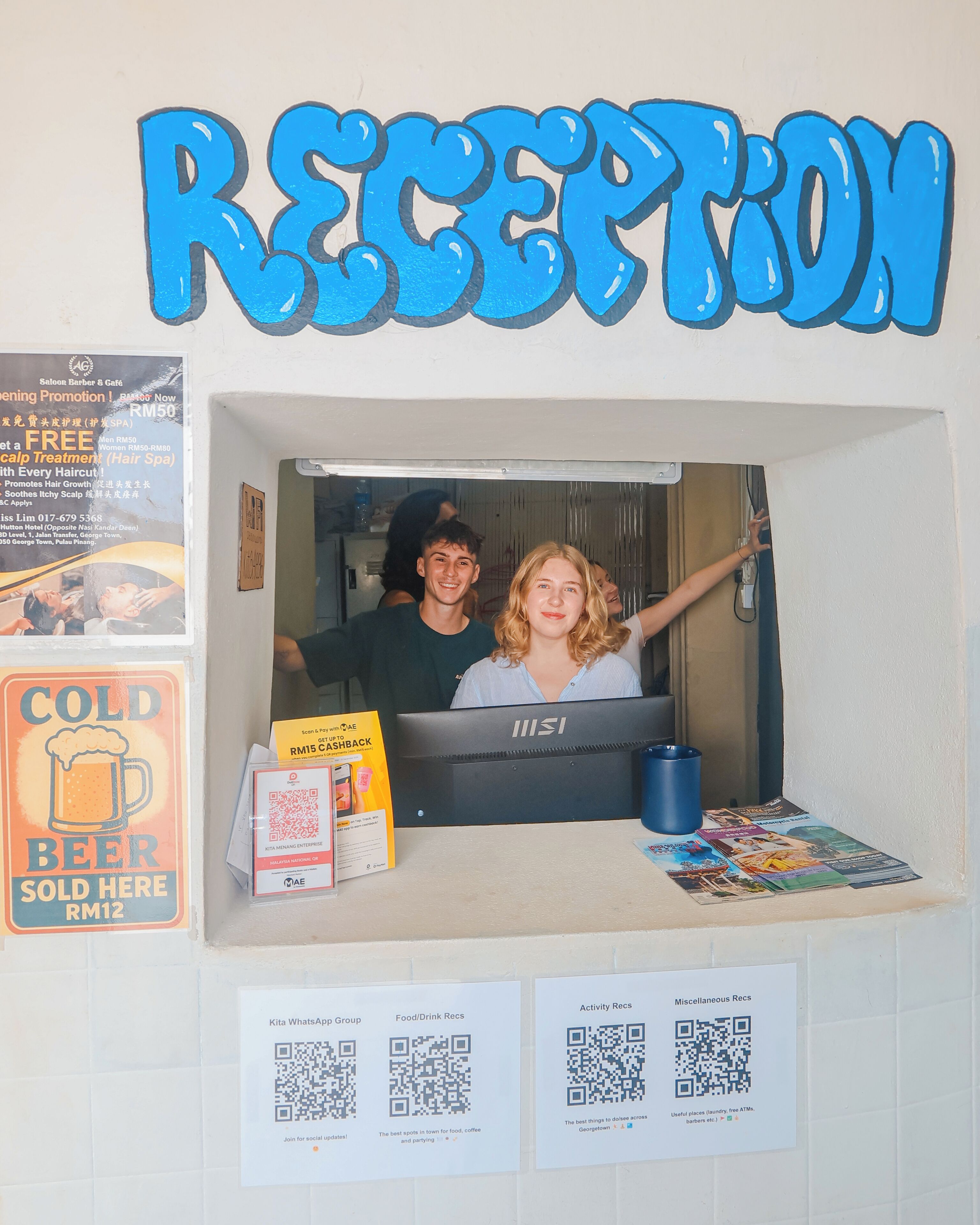 A reception area with a sign reading "RECEPTION" above a counter. There are promotional posters on the wall, a QR code for a mobile app, and a display of a cold beer for RM12.
