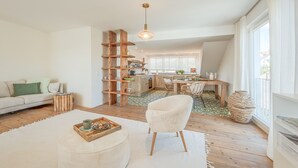 Ein modernes Wohnzimmer mit einer Couch, einem Hocker und einem weißen Sessel. Im Hintergrund ist ein Bücherregal aus Holz und ein Essbereich mit Tisch und Stühlen zu sehen.