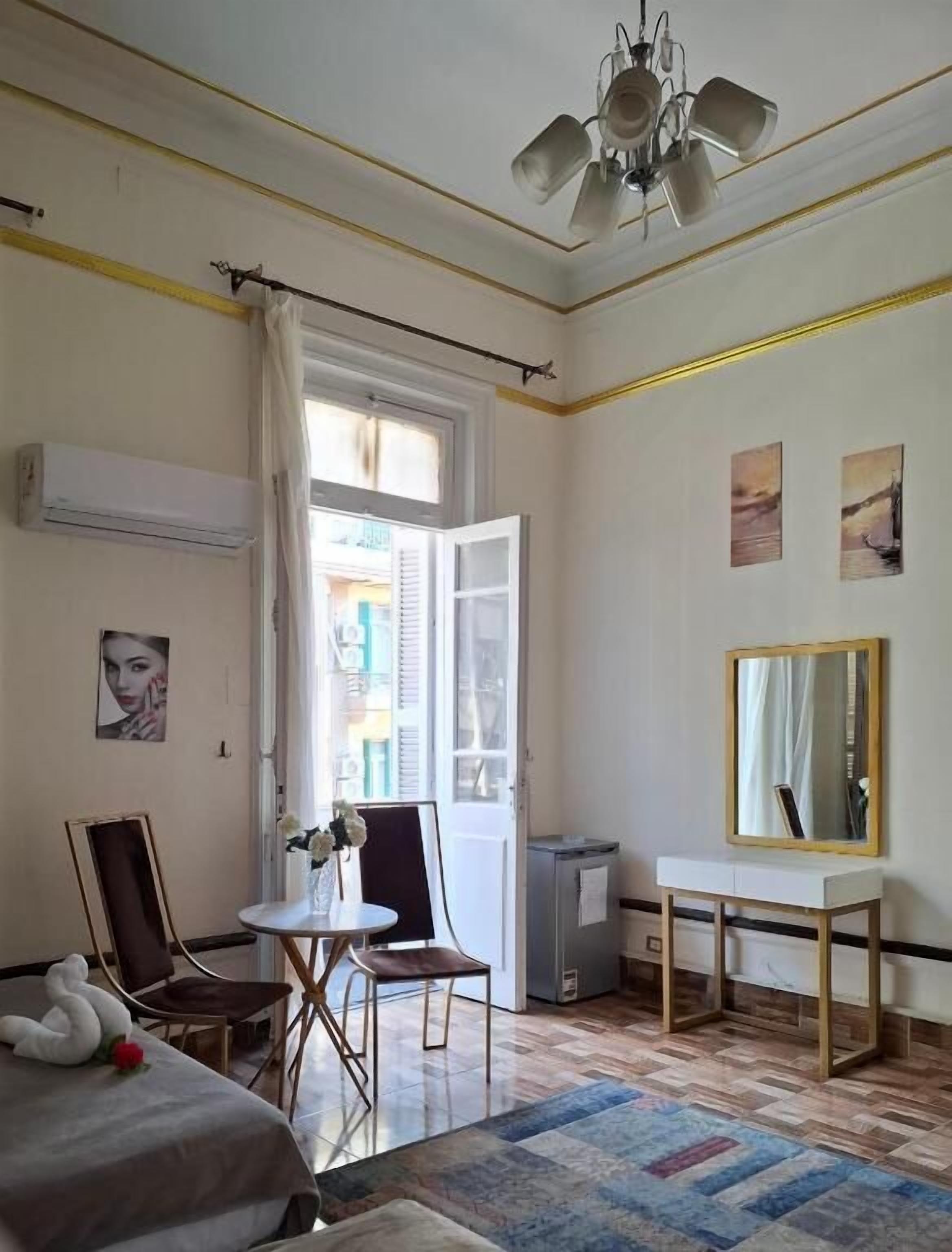 Una camera d'albergo con un letto, una scrivania, una sedia, un tavolino con fiori, una ventola a soffitto e una finestra con tende.