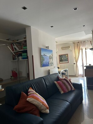 A living room with a dark blue sofa, colorful pillows, a painting of a landscape, a dining area with a table and chairs, and a staircase leading to another level. - Ketty's House Apartment between sea and mountains (Vico Equense)