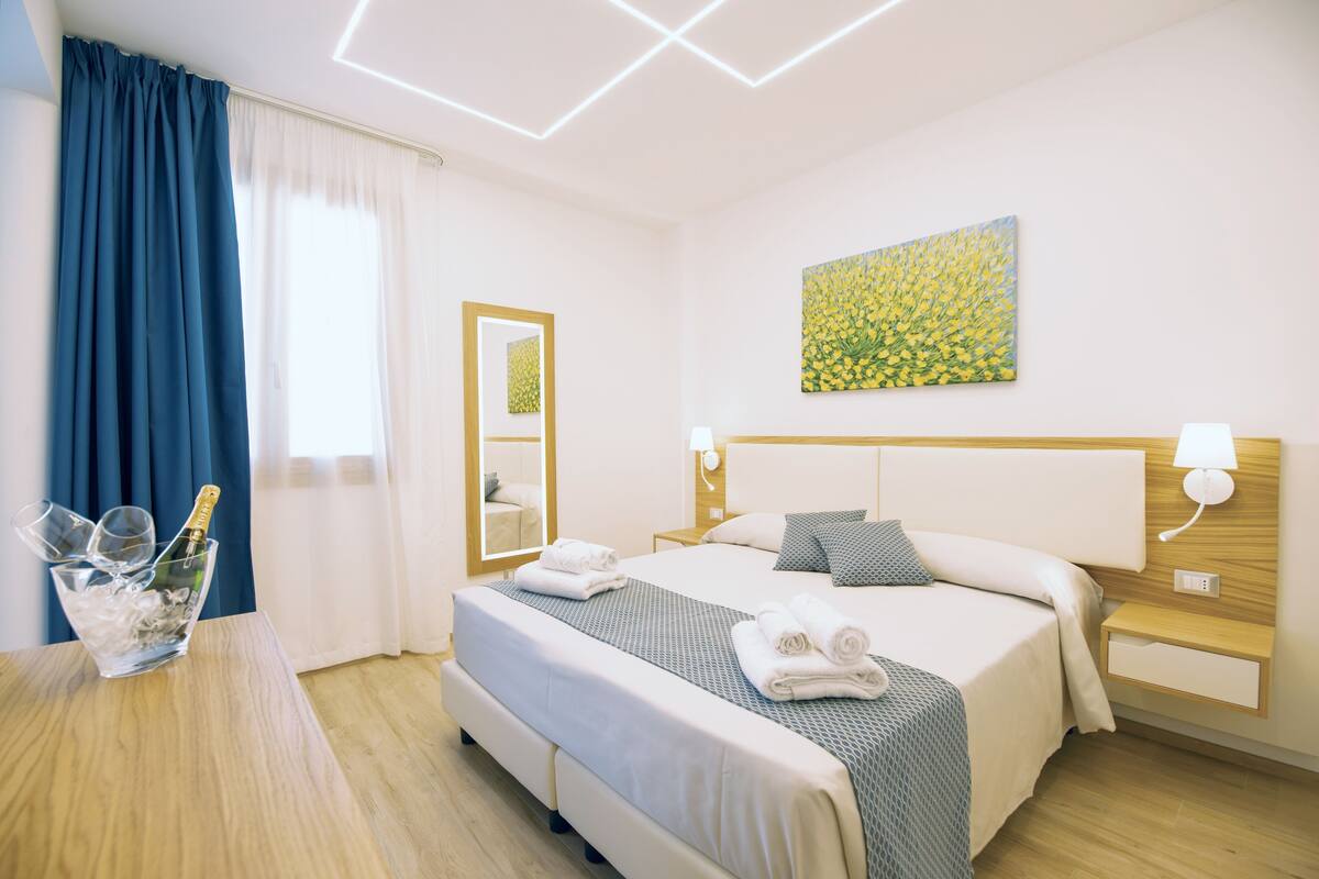Una habitación de hotel con una cama, mesitas de noche, un espejo, un cuadro y una ventana con cortinas.