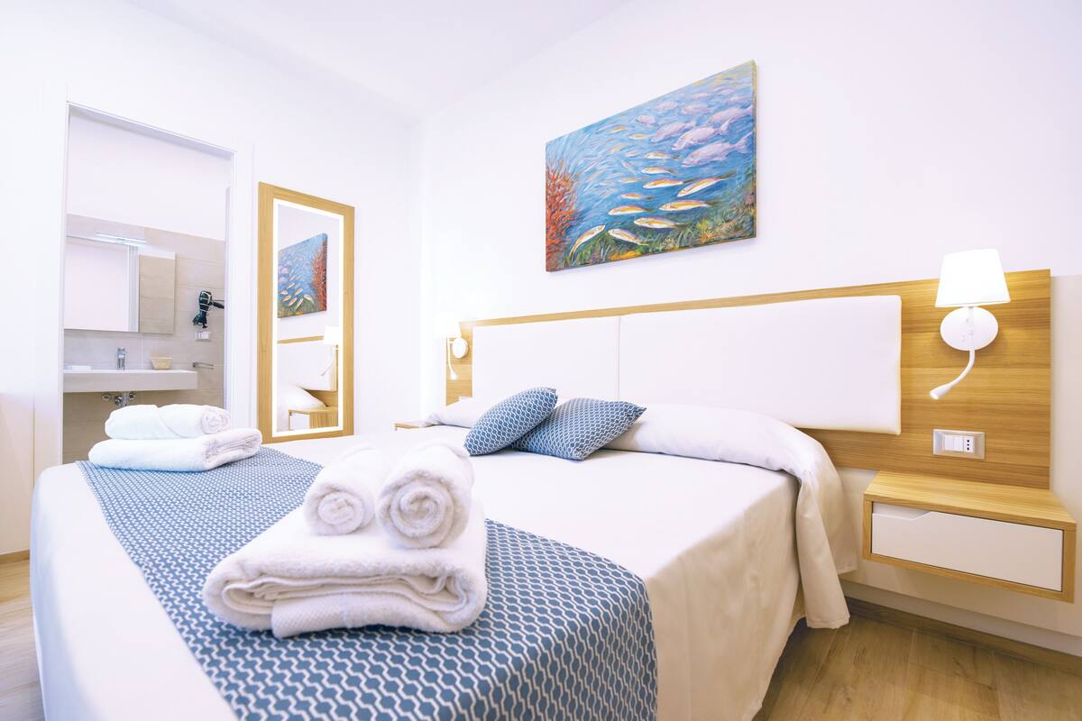 Habitación de hotel con cama, mesita de noche, lámpara de pared, un cuadro, espejo y un baño visible a través de una puerta abierta.