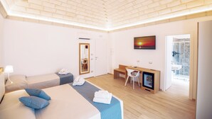 A modern hotel room with a brick-patterned ceiling, a large bed with blue patterned pillows, a desk with a chair, a television, and a bathroom visible through an open door. - I Mori Hotel  (San Vito Lo Capo)