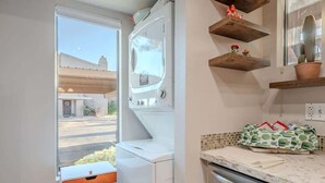 A compact kitchen with a built-in washer and dryer, a window with a view of the street, and wooden shelves with potted plants. - Starlight Escape - Heated Pool & Spa by Old Town! (Scottsdale)
