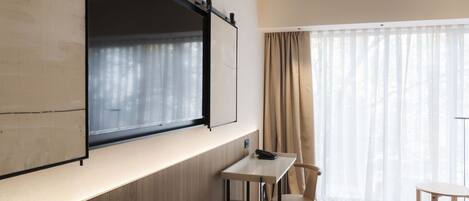 A modern room with a large window, a flat-screen TV mounted on the wall, a small table, and a chair.